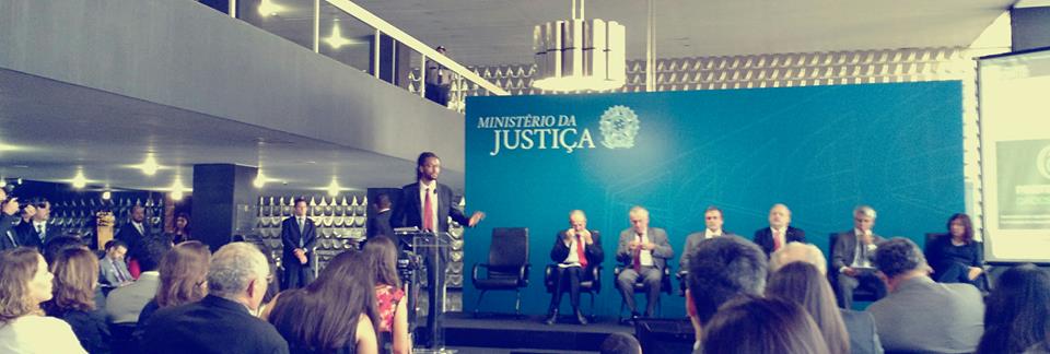 Photo of a speech given at the Ministry of Justice of Brazil, where a man presents himself speaking in front of a platform, with four authorities seated behind him, in front of a blue sign with the phrase "Ministry of Justice" and public seated in front of him .