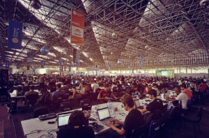 Imagem de um galpão com muitas mesas longas, brancas e compartilhadas. Nas mesas, as pessoas estão sentadas usando computadores.
