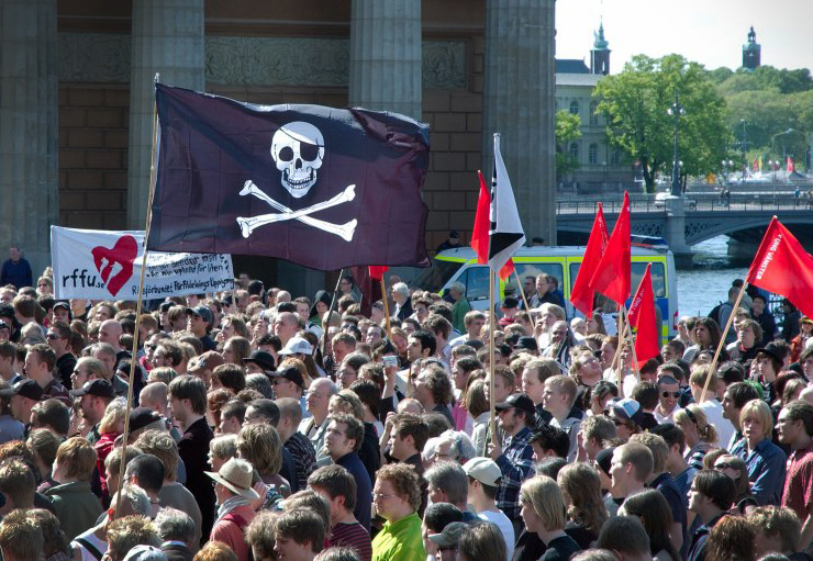 Photo of white people protesting. Some hold red flags. In the central part of the image, there is a black flag with a skull, referring to pirate flags.