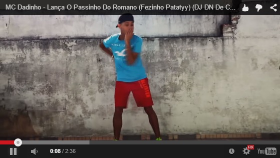 PrintScreen de um vídeo do YouTube. No Vídeo, um menino negro, de bermuda vermelha, camiseta azul, e boné preto está dançando. A legenda do vídeo é "MC Dadinho - passinho do romano".