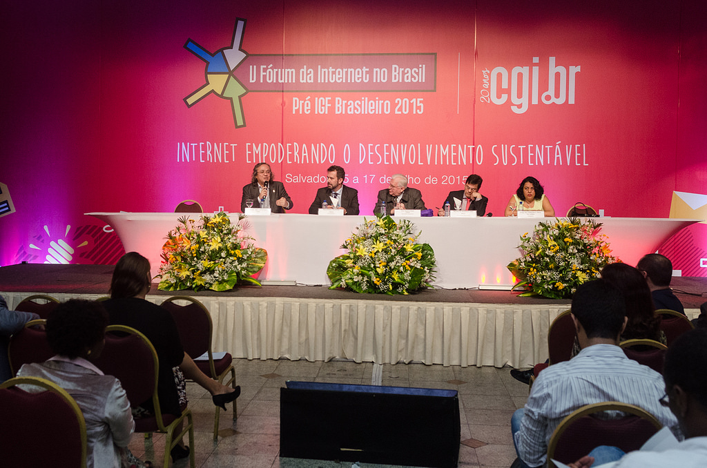 Photo of the Internet Forum, where five people are seated at the event's board of directors, in front of a pink panel with the logos of the event "V Fórum da Internet no Brasil: Pré IGF Brasileiro 2015" and cgi.br, and the title of the event"Internet empoderando o desenvolvimento sustentável". In front of the table, three flower ornaments are arranged and the audience is seated in rows.