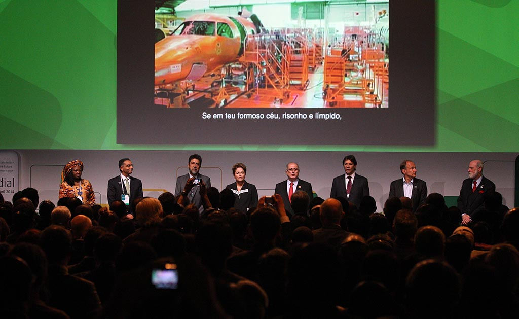 Photo of the NETMundial event, with 8 authorities siting on a stage, among them the President of the Republic Dilma Rousseff and mayor of the city of São Paulo Fernando Haddad, sitting in front of a screen showing the image of an industry with the passage "Se em teu formoso céu, risonho e límpido"  of the Brazilian National Anthem, in front of the audience formed by several people standing.