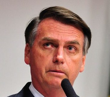 Imagem do então pré-candidato à presidência Jair Bolsonaro, com semblante sério, vestindo terno e gravata e com um microfone à sua frente.