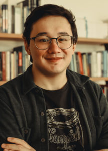 Foto de André Houang, homem asiático de cabelos pretos. Aparece sorrindo de óculos de grau e roupas pretas. No fundo, aparecem livros. Foto de Bianca Moreira/InternetLab.