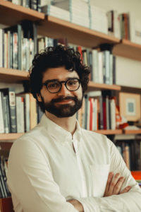 Foto de Artur Pericles, homem branco, de óculos de grau, barba média e cabelos pretos. Veste uma blusa branca de mangas longas e está com os braços cruzados. Aparece sorrindo. Ao fundo, uma estante de livros. Créditos da imagem: Bianca Moreira/InternetLab.