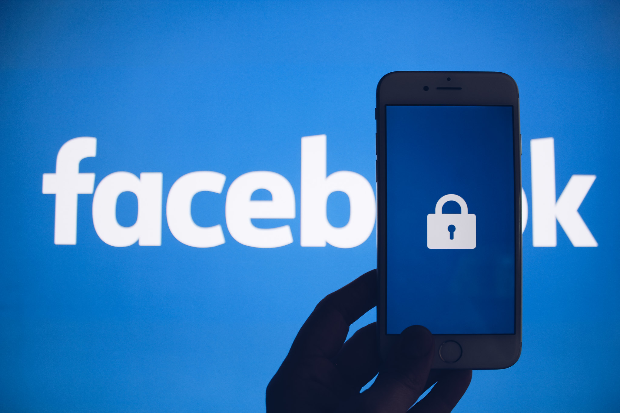 Image on a blue background which says "Facebook" with a hand holding a smartphone in front