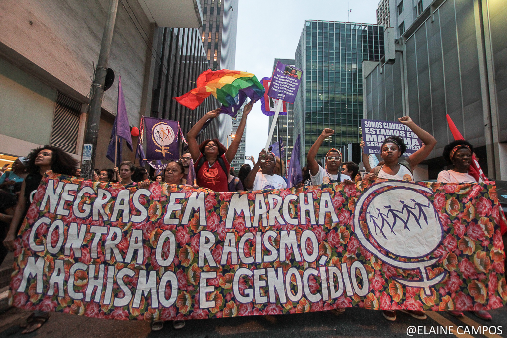 Imagem mostra a Marcha das Mulheres Negras, com mulheres segurando bandaeira LGBTQIA+ e uma faixa escrito "Negras em marcha contra o racismo, machismo e genocídio".